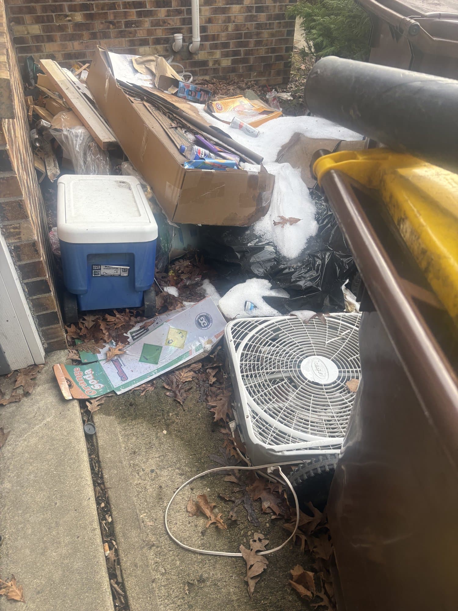 Pile of household junk — cooler, boxes, fan, and bagged debris stacked against a brick wall