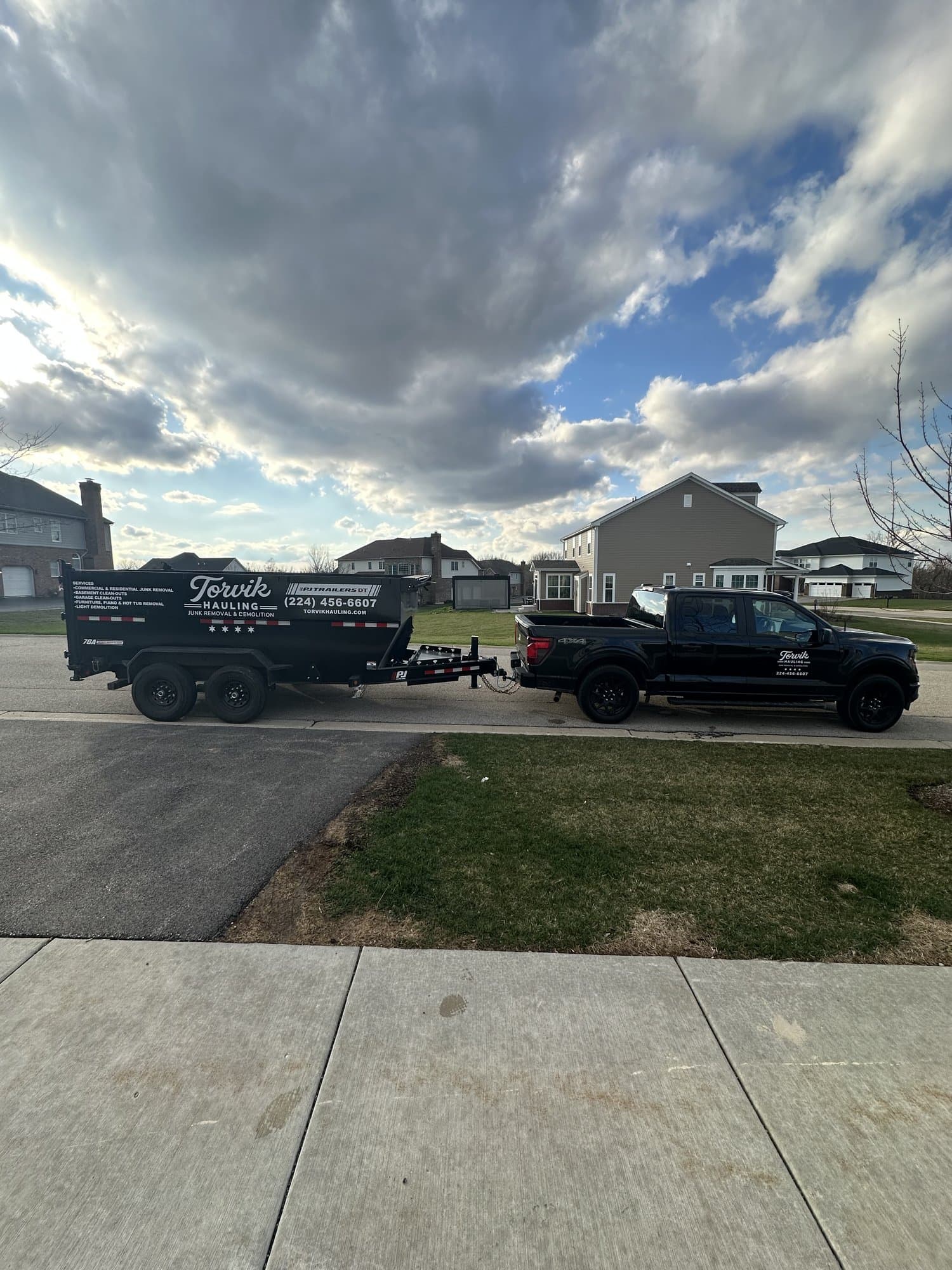Branded Torvik Hauling pickup truck pulling the dump trailer through a Northwest Chicago suburb under a dramatic cloudy sky