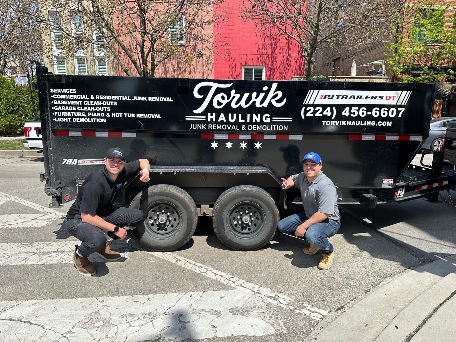 The Torvik Hauling team kneeling beside their branded dump trailer with services list and phone number on the panel