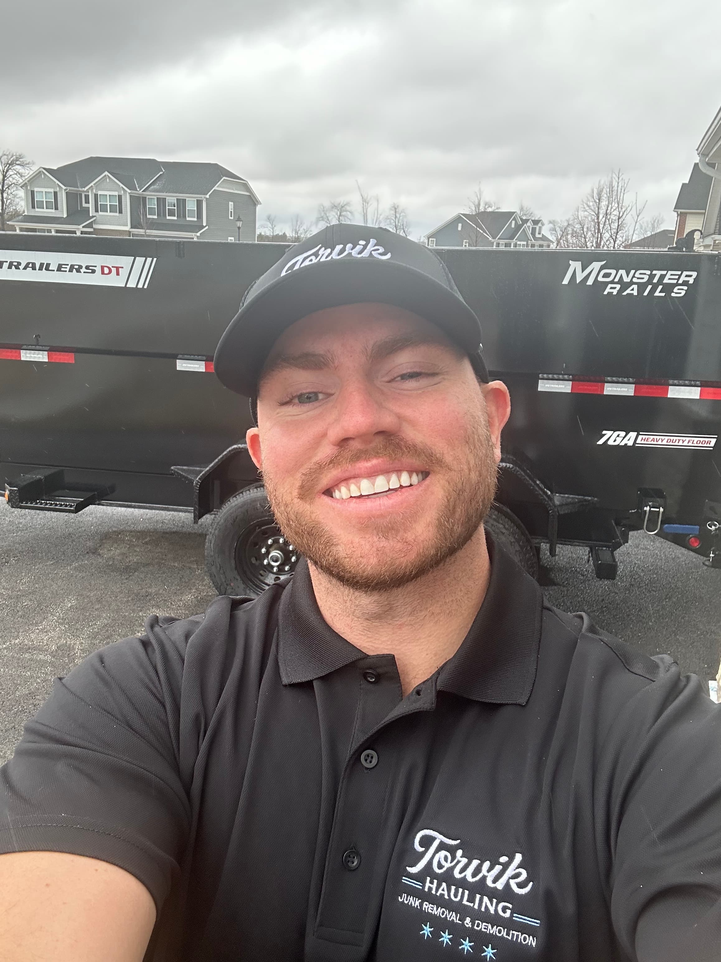 Torvik Hauling owner smiling in a branded black Torvik polo and cap beside the Torvik dump trailer